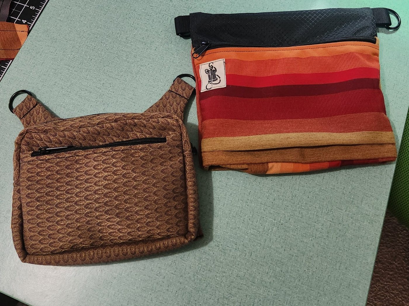 Two crossbody bags on a sewing table: one in woven brown fabric with a front zip pocket, and one in orange and red stripes with a black top panel