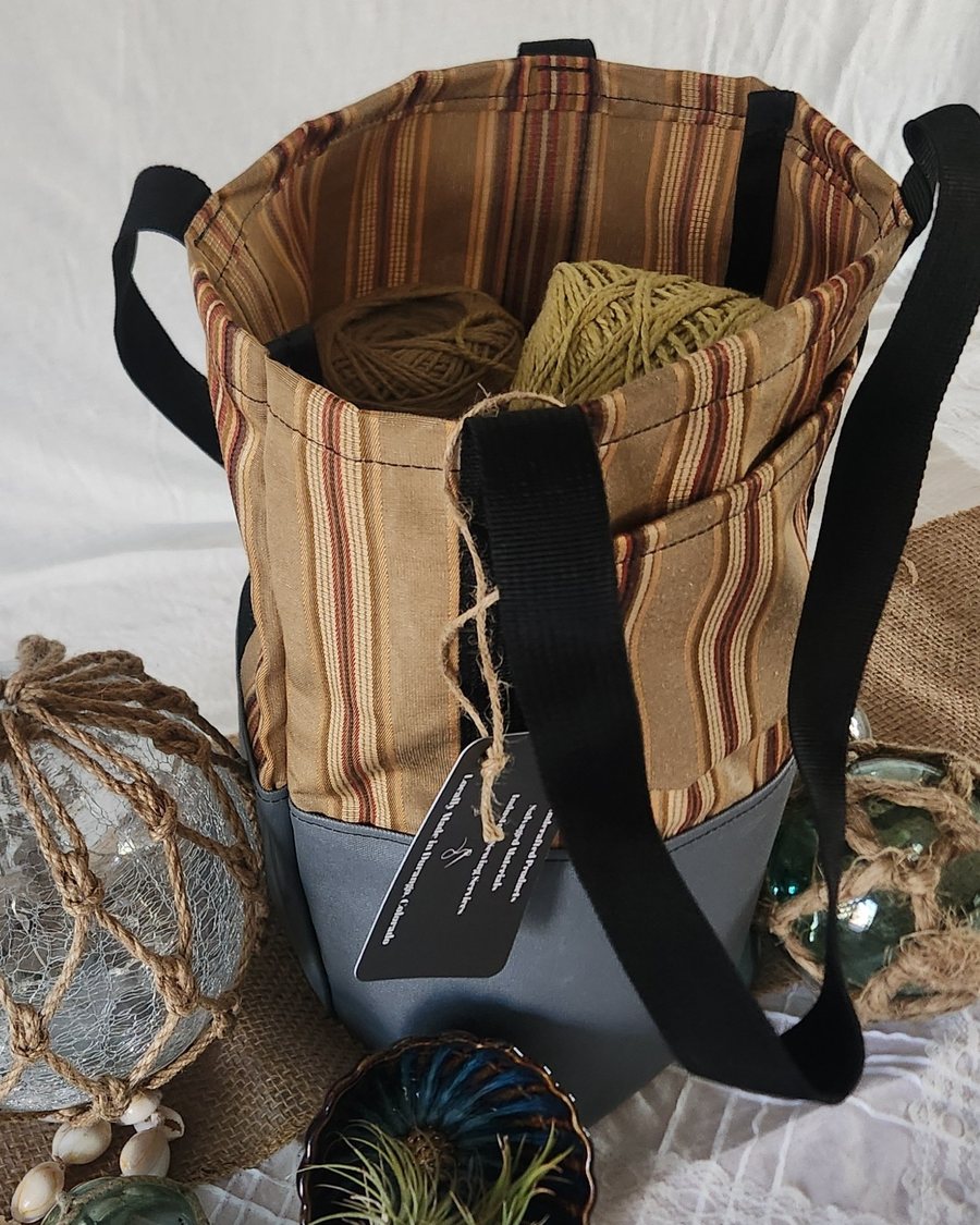 Small striped tote with a gray base and black webbing handles, holding two balls of yarn, shown with glass fishing floats and shells