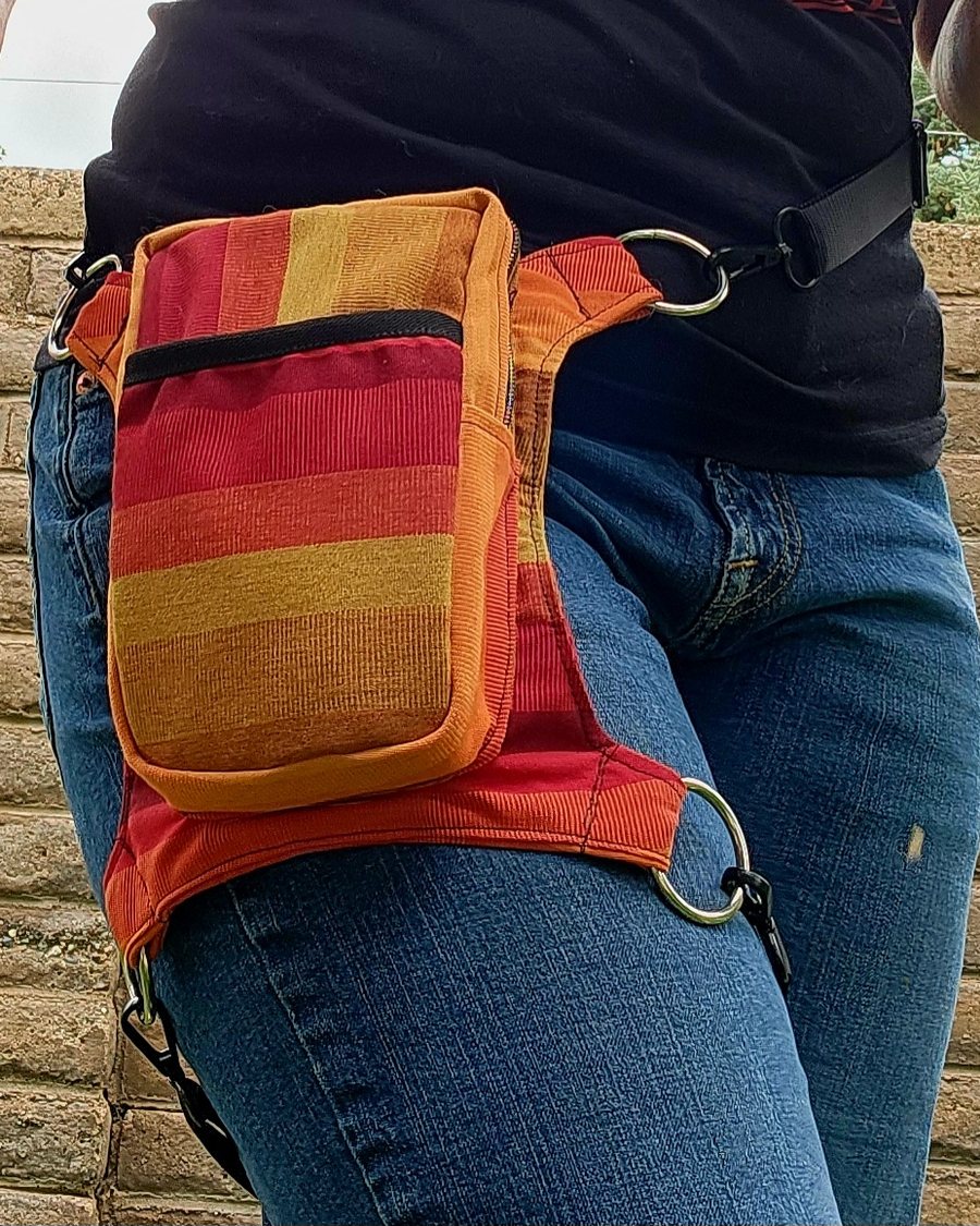 Hip bag in orange, red, and gold stripes with a front zip pocket, metal rings, and a thigh strap, worn at the hip