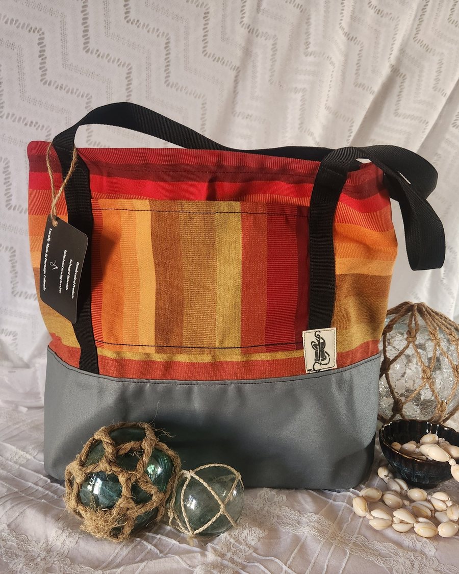 Tote bag with red, orange, and gold stripes above a gray base, black webbing handles, and a hang tag, shown with glass fishing floats and cowrie shells